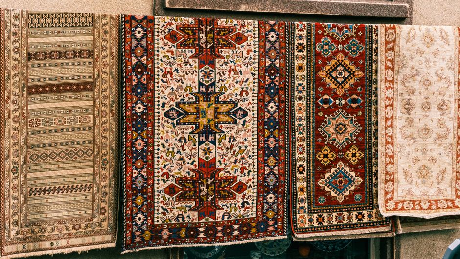 A close-up view of four intricately patterned Persian and Oriental rugs displayed side by side on a flat surface, showcasing their detailed geometric and floral motifs in various colors including red,